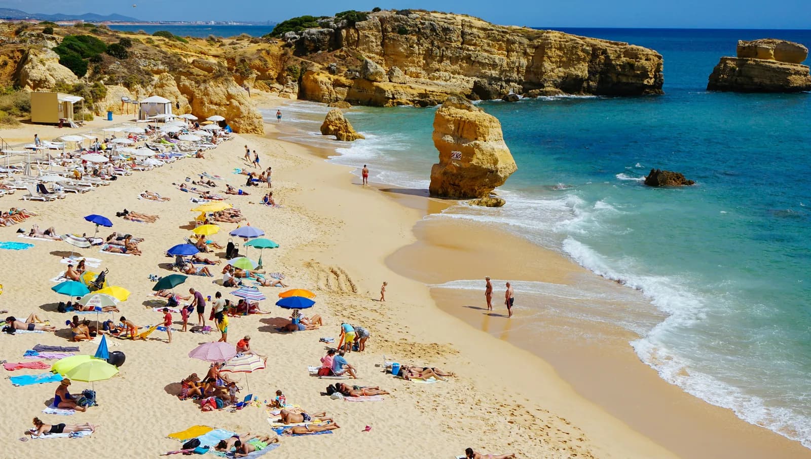 A busy Algarve cove with sun loungers below golden sandstone cliffs.